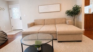 A living room with a beige sectional sofa, a glass coffee table with a plant, a framed artwork on the wall, and a wooden cabinet in the background. - NYC Bus at Door • EWR • Outlet • MetLife • Parking - 3-bedroom apart, 1st floor. (Elizabeth)