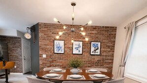 A dining area with a brick wall, a chandelier, and framed cityscape pictures. - NYC Bus at Door • EWR • Outlet • MetLife • Parking - 3-bedroom apart, 1st floor. (Elizabeth)