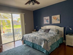 Ein Schlafzimmer mit Bett, Nachttisch, Lampe, Deckenventilator und Blick auf einen Pool sowie Sitzgelegenheiten im Freien.