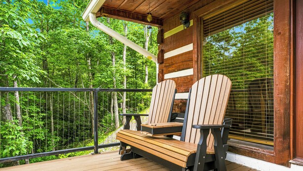 Ett trädäck med Adirondack-stolar, ett räcke och ett fönster med persienner, med utsikt över en skog.