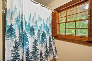 A bathroom with a shower curtain featuring a forest print, a window with wooden shutters, and a view of greenery outside. - Mountain Views, Hot Tub, Pool Access (Gatlinburg)