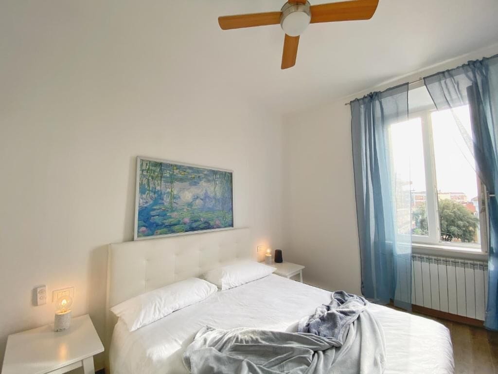 Un dormitorio con cama, ventilador de techo, un cuadro, mesita de noche, lámpara y ventana con cortinas.