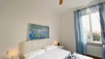 Un dormitorio con cama, ventilador de techo, un cuadro, mesita de noche, lámpara y ventana con cortinas.