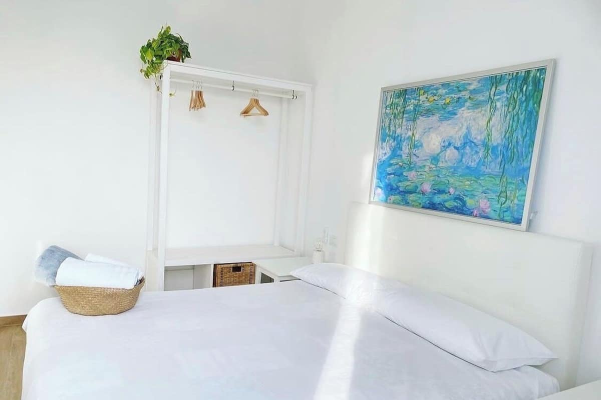 A bedroom with a bed, a wall-mounted painting, a hanging plant, and a basket with towels.
