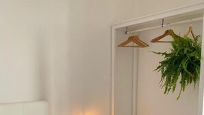 A bedroom with a bed, bedside table with a candle, a hanging plant, and a white wardrobe with shelves.