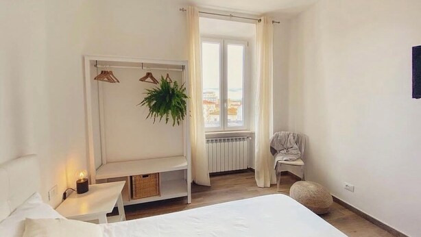 A bedroom with a bed, a bedside table with a lamp, a hanging plant, a radiator, a window with curtains, and a chair.