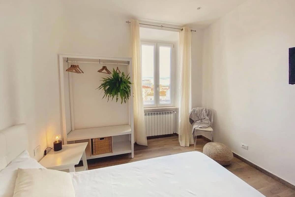 A bedroom with a bed, a bedside table with a lamp, a hanging plant, a radiator, a window with curtains, and a chair.