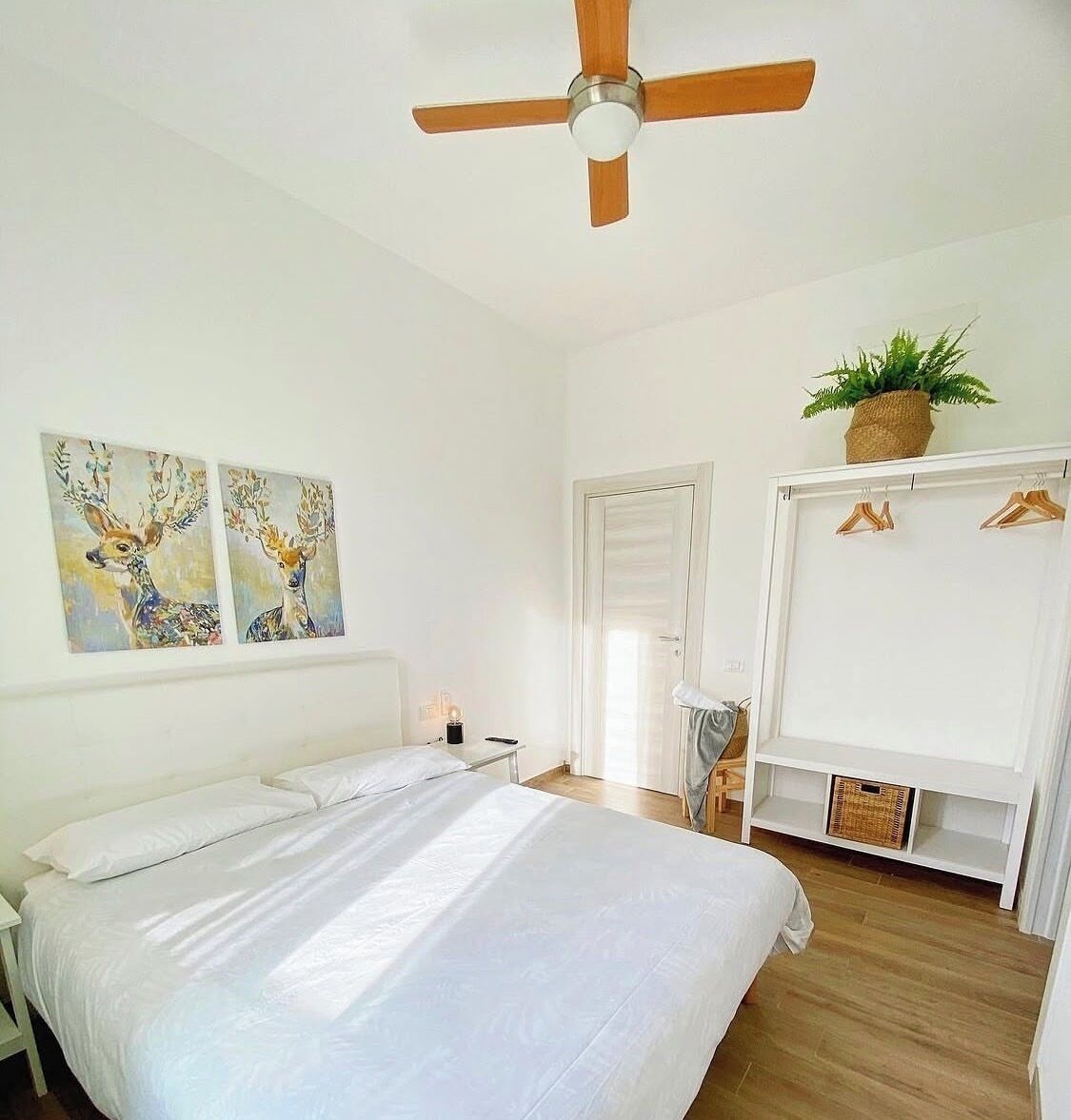 Une chambre à coucher comprenant un lit, un ventilateur de plafond, une porte, une étagère fixée au mur avec une plante, et deux œuvres d’art mural.