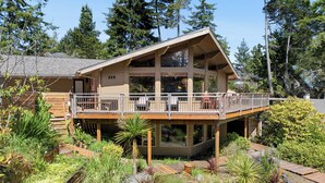 A two-story house with a balcony, surrounded by a garden with various plants and rocks. - Lodge style 5 bedroom, 2 kitchens, large deck, outdoor firepit, 1 dog ok (Manzanita)