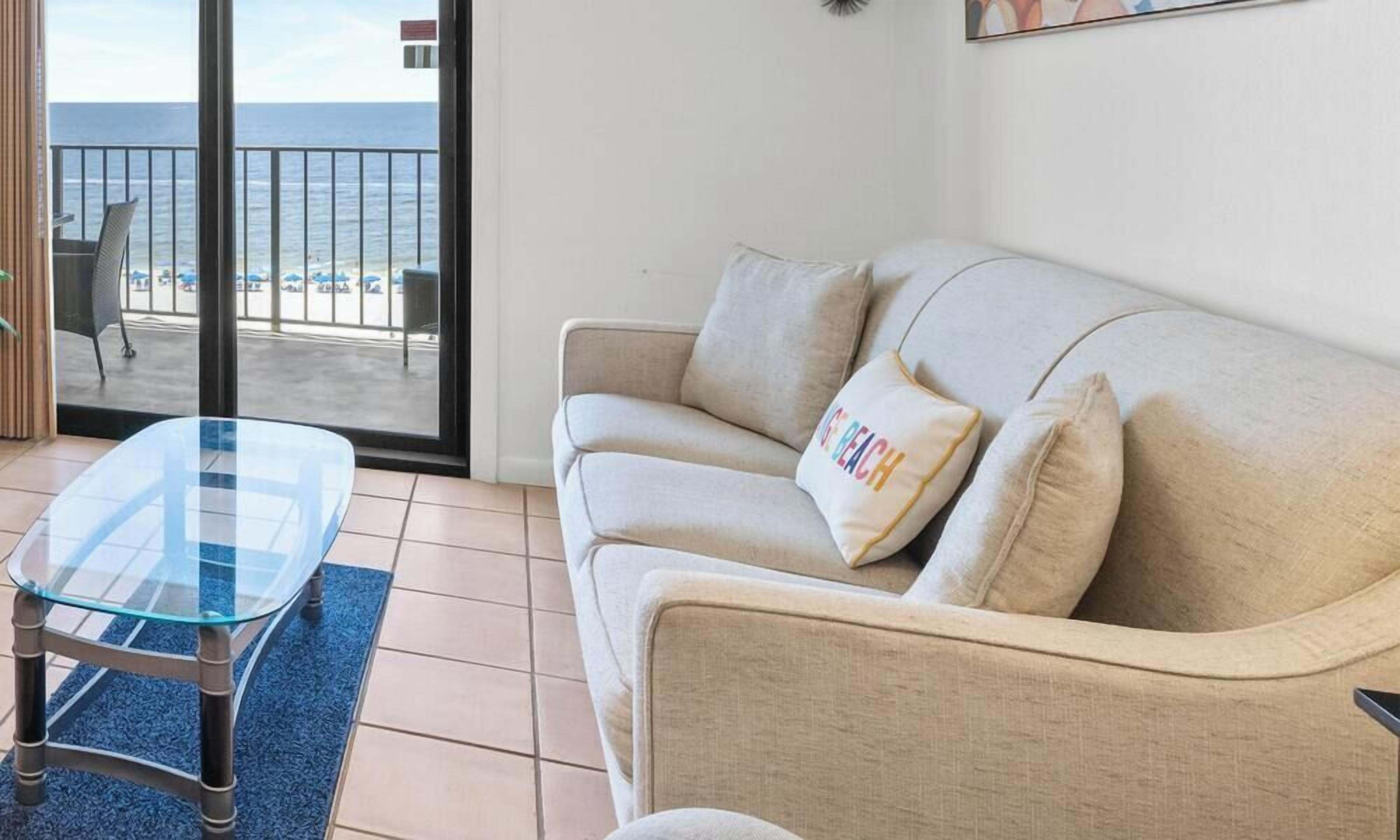 Un salon avec un canapé, une table basse en verre et une vue sur la plage, accessible par une porte coulissante.