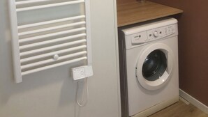 Une buanderie avec un sèche-linge blanc fixé au mur, une étagère en bois et un radiateur blanc.