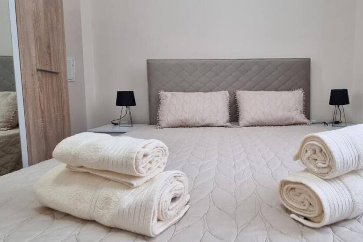 Una cama bien hecha con toallas blancas enrolladas, un cabecero, dos lámparas de noche y un espejo a la izquierda.
