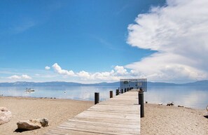 Eine Holzstege führt zu einem Steg mit Metallgeländer, einem Sandstrand und ruhigem Wasser, im Hintergrund Berge.