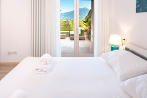 A hotel room with a bed, a nightstand, a lamp, and a view of a balcony with outdoor furniture and a mountain in the background. - 640A Sonja's House (Torri del Benaco)