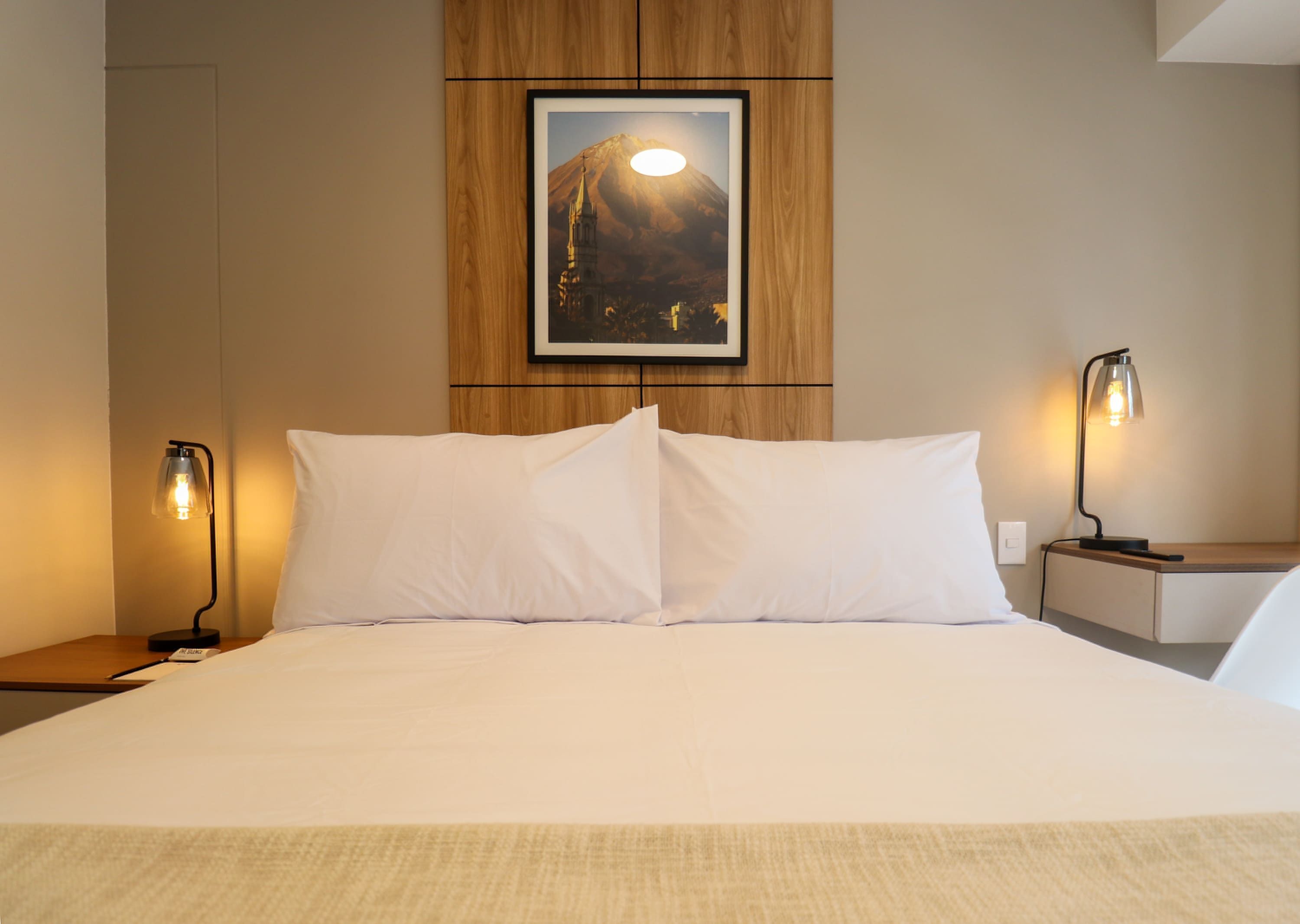 A neatly made bed with white linens, two bedside lamps, a framed picture of a mountain, and a wooden panel wall.