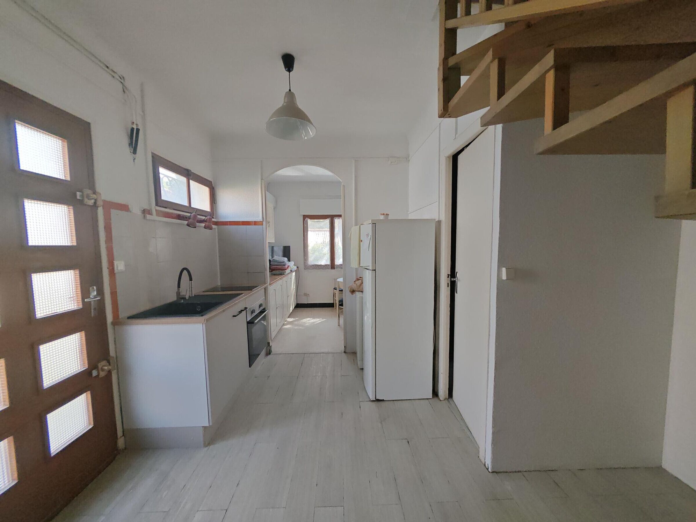 A narrow hallway with a kitchen area on one side, a wooden staircase, and a door leading to another room.