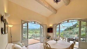 A room with a dining table, chairs, a sofa, and a view through large windows.