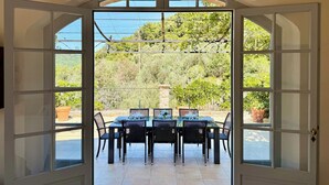 An open door leading to a patio with a table and chairs, surrounded by potted plants and a view of greenery.