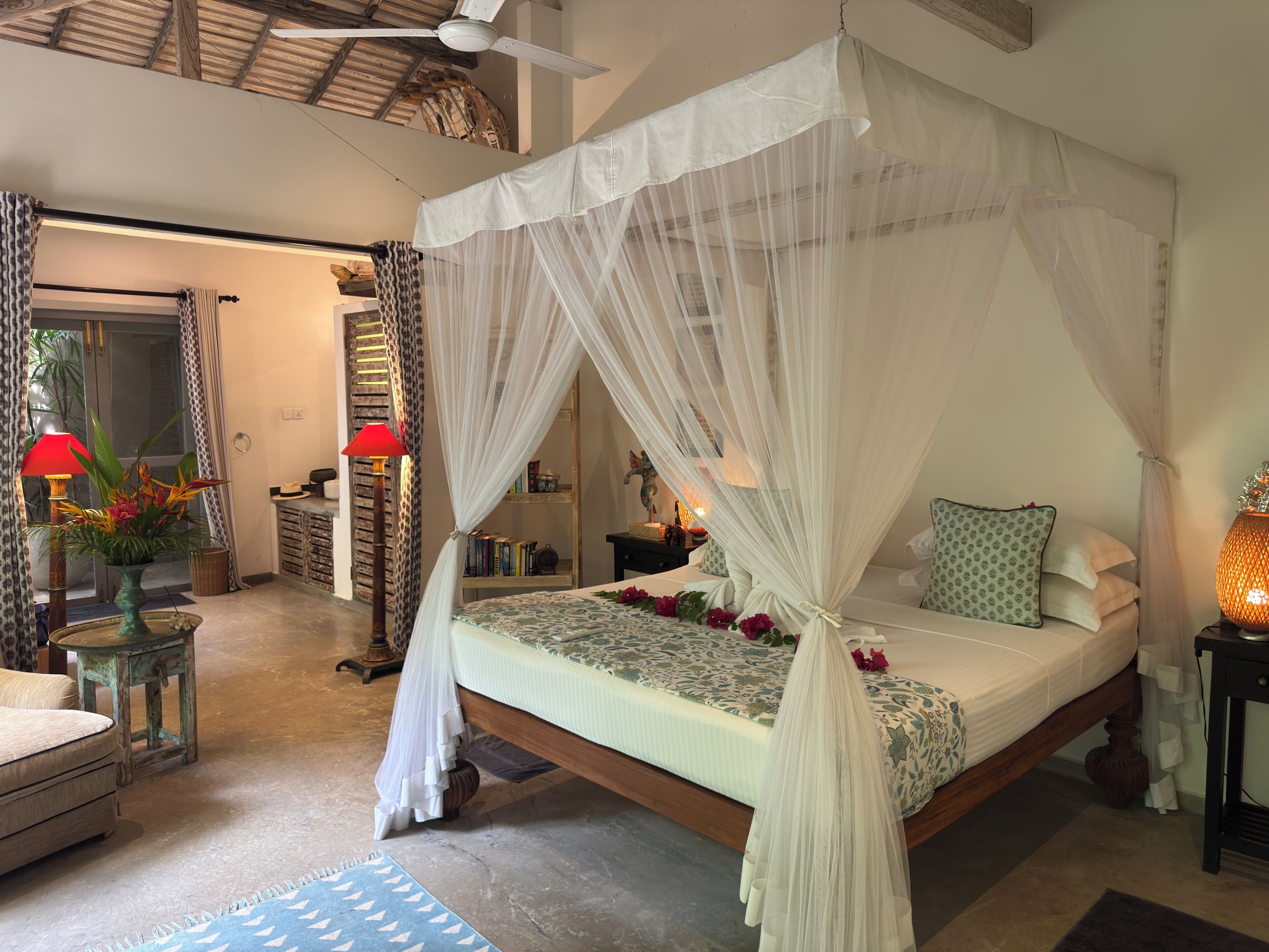 A bedroom with a canopy bed, a bedside table with a lamp, a nightstand, and a decorative vase with flowers.