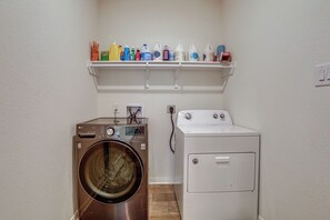 A laundry room with a front-loading washer and dryer, a shelf with cleaning supplies, and a wall-mounted electrical outlet.