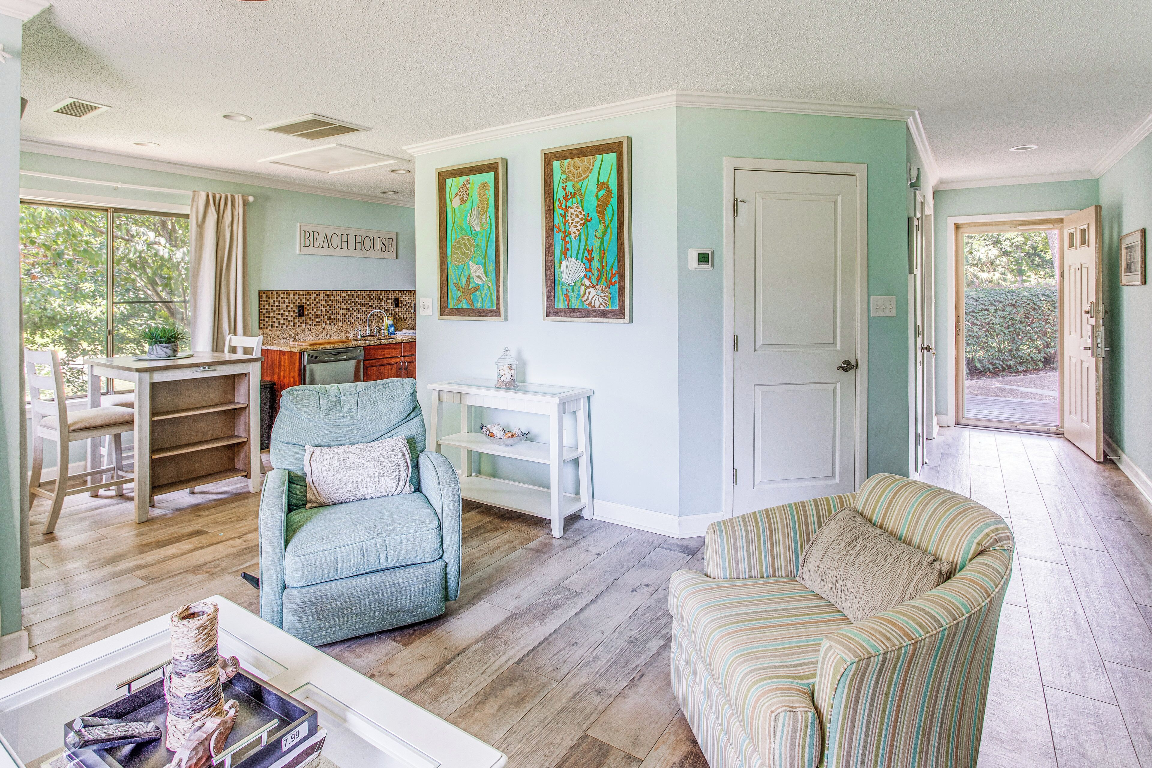 A living room with a sofa, a chair, a dining table with chairs, a kitchen area, and a door leading outside.