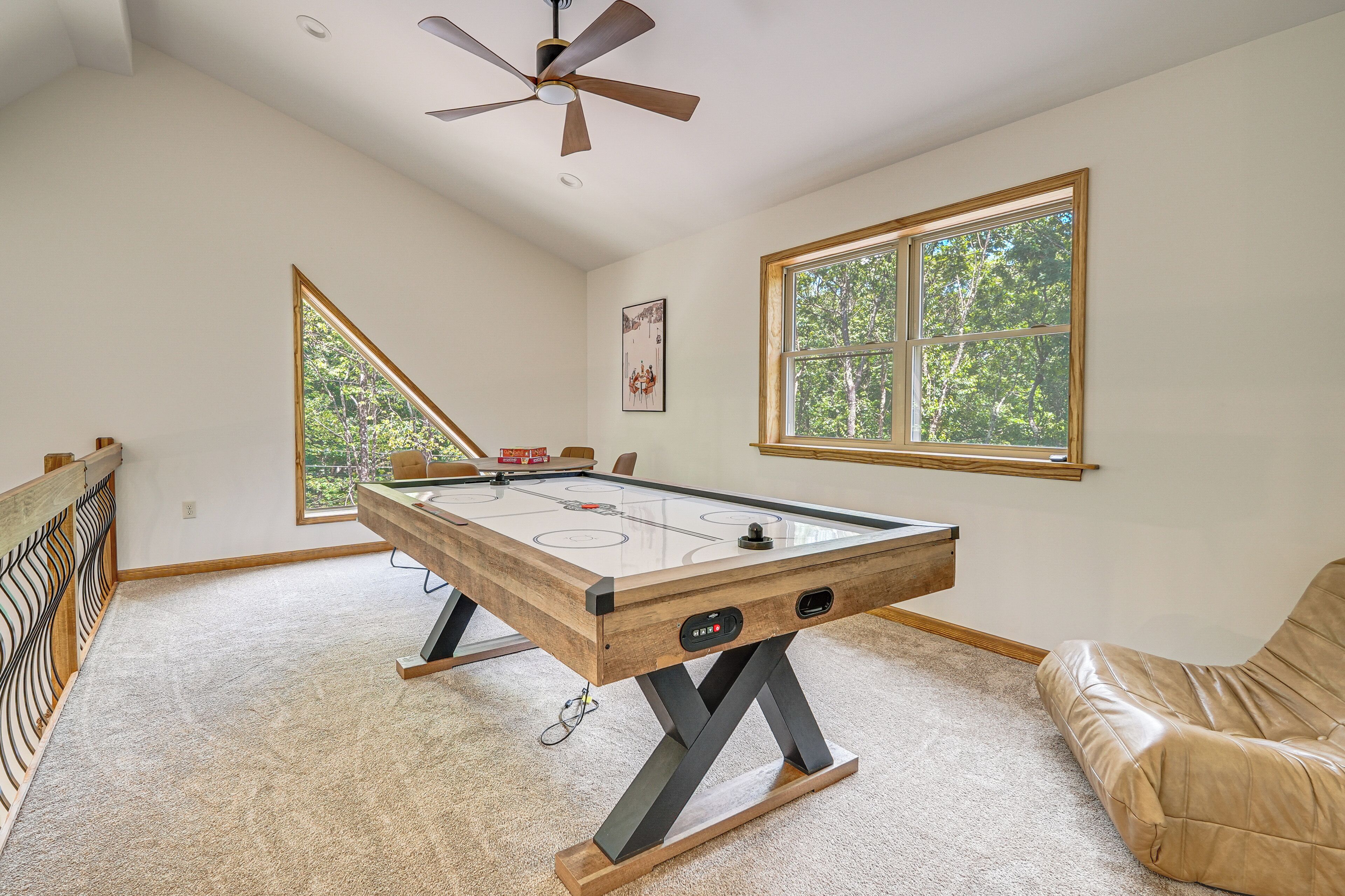 A room with a pool table, a ceiling fan, a window with a view of trees, and a brown leather couch.