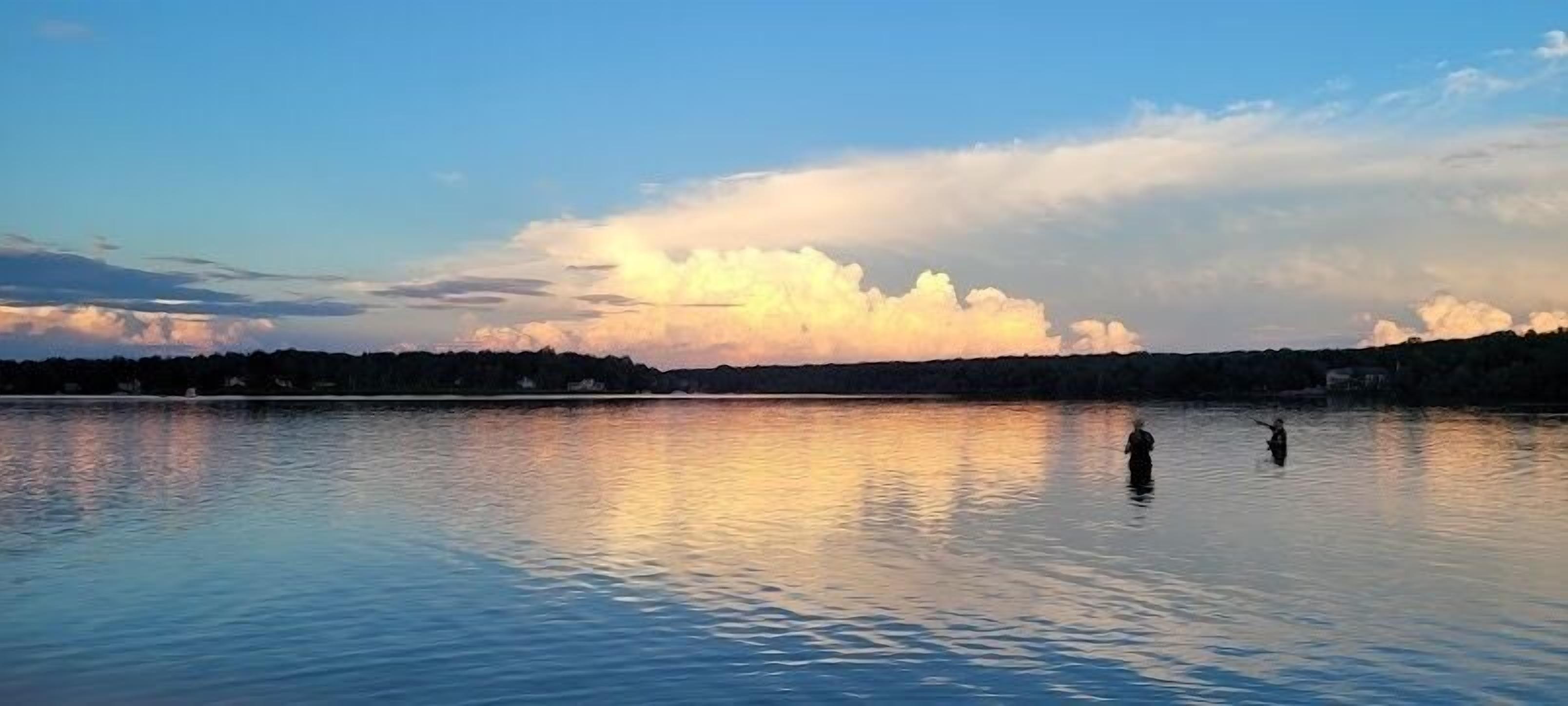 Un lac paisible où se tiennent deux personnes dans l’eau, entourés d’une rive bordée d’arbres.
