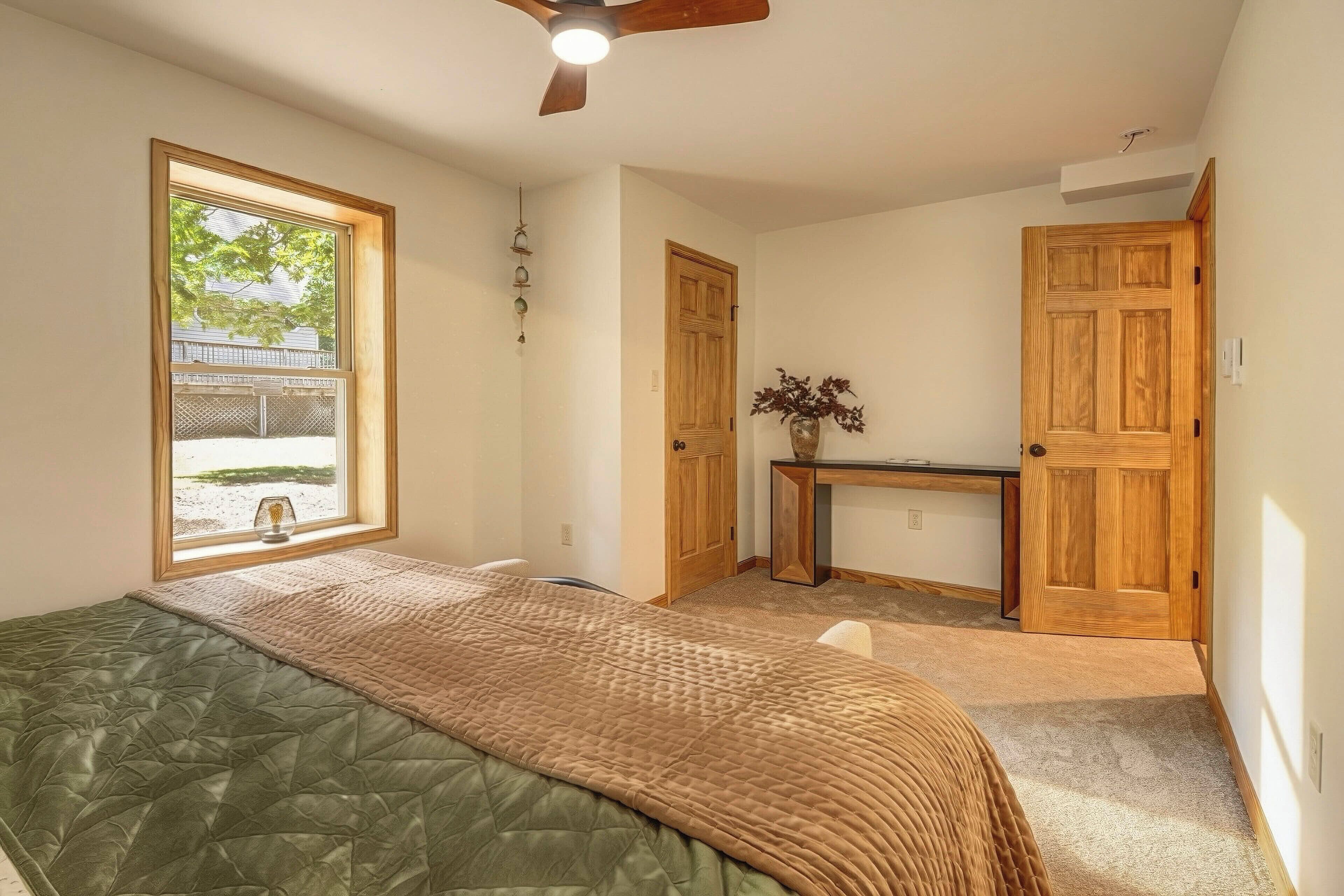 A bedroom with a bed, a desk, and a window with a view of a fence and trees.