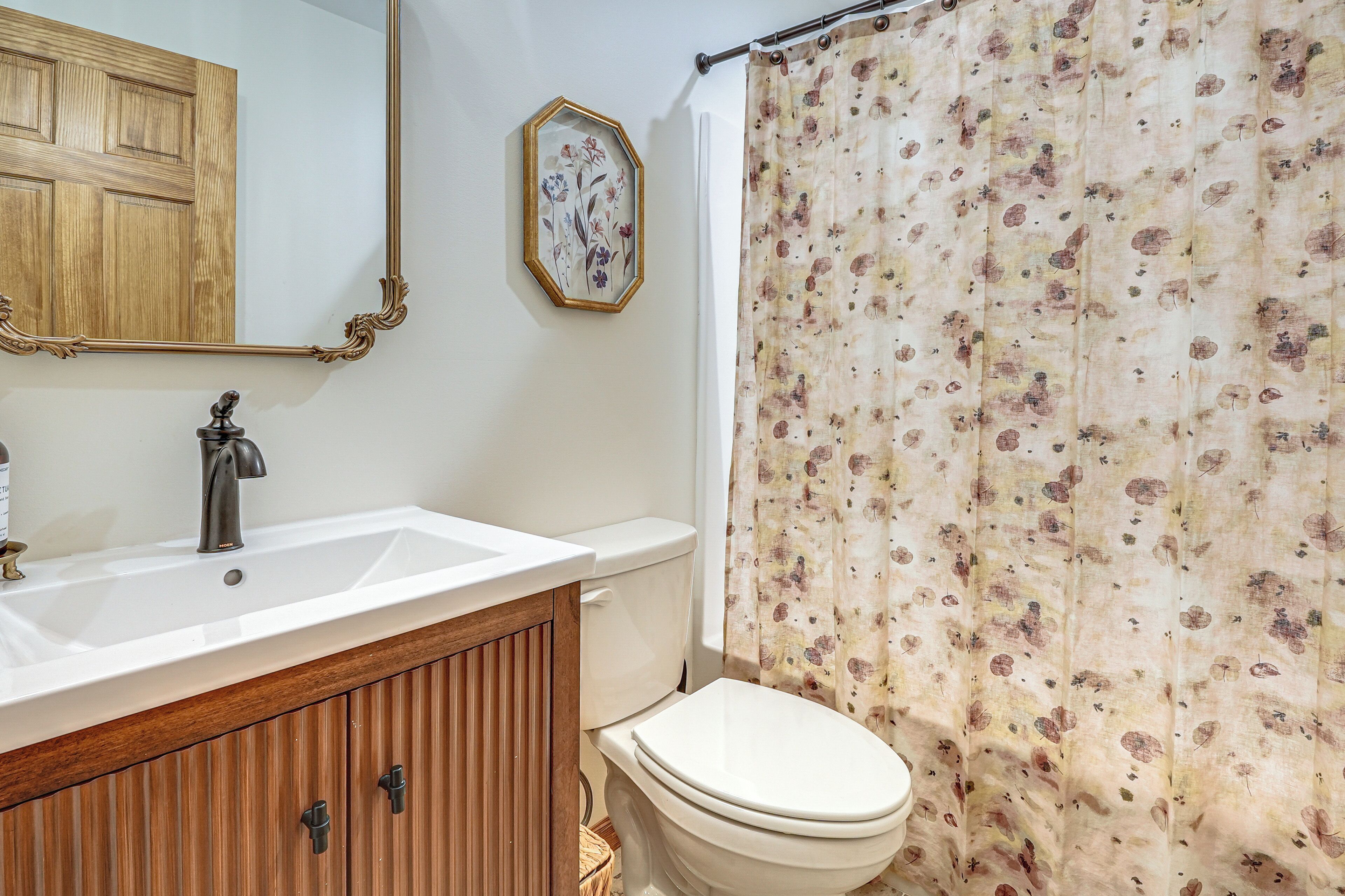 A bathroom with a wooden vanity, a white sink, a toilet, a shower curtain with a floral pattern, a mirror with an ornate frame, and a wooden door.