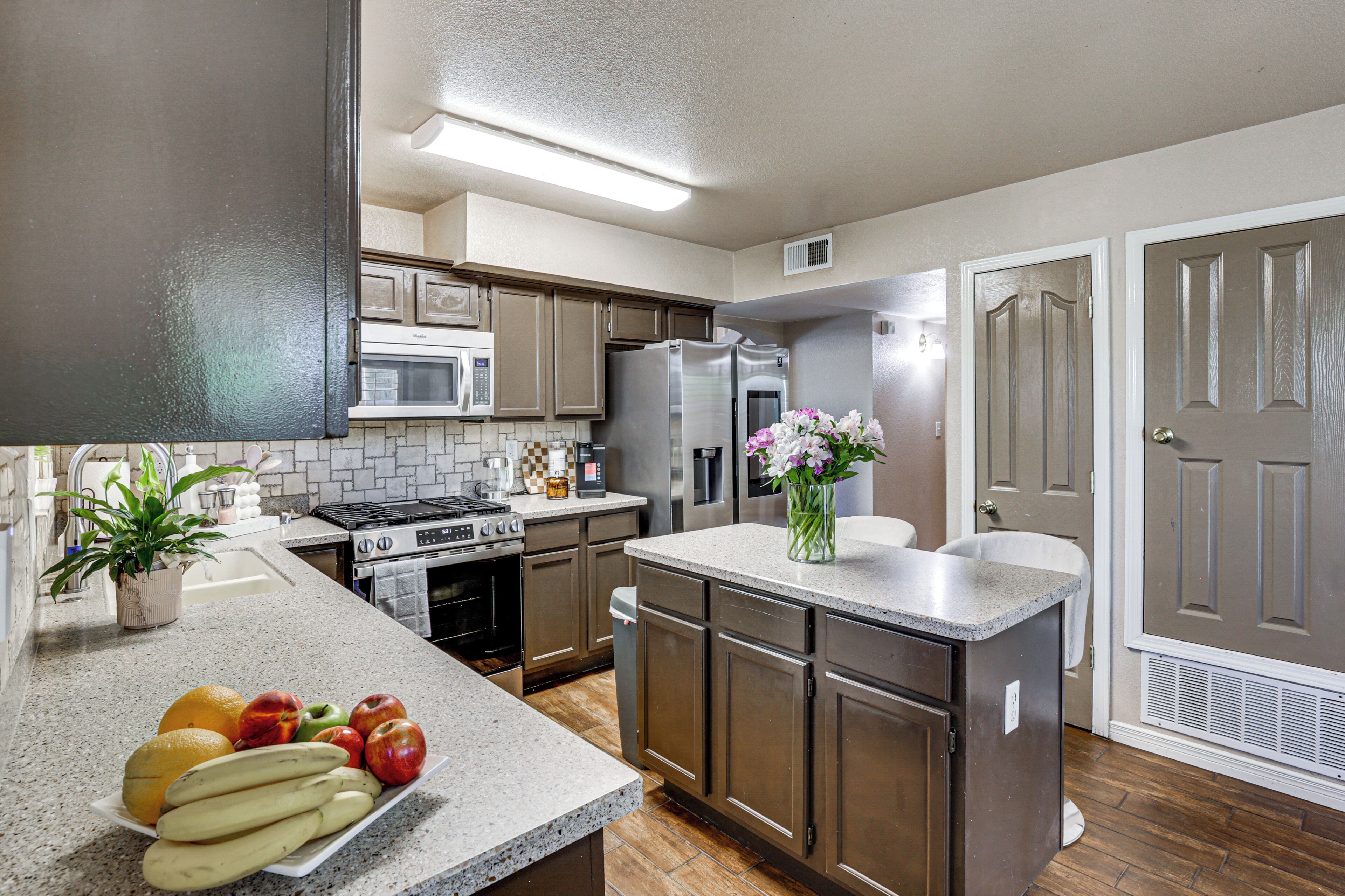 A kitchen with a stove, microwave, refrigerator, and a sink. There is a fruit bowl with bananas and apples, and a vase with flowers on the counter.