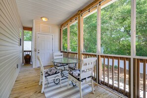 A covered porch with a glass table and wicker chairs, a wooden deck, and a view of trees. - Golf Cart, Bikes, Easy Beach Days! Oak Island (Oak Island)