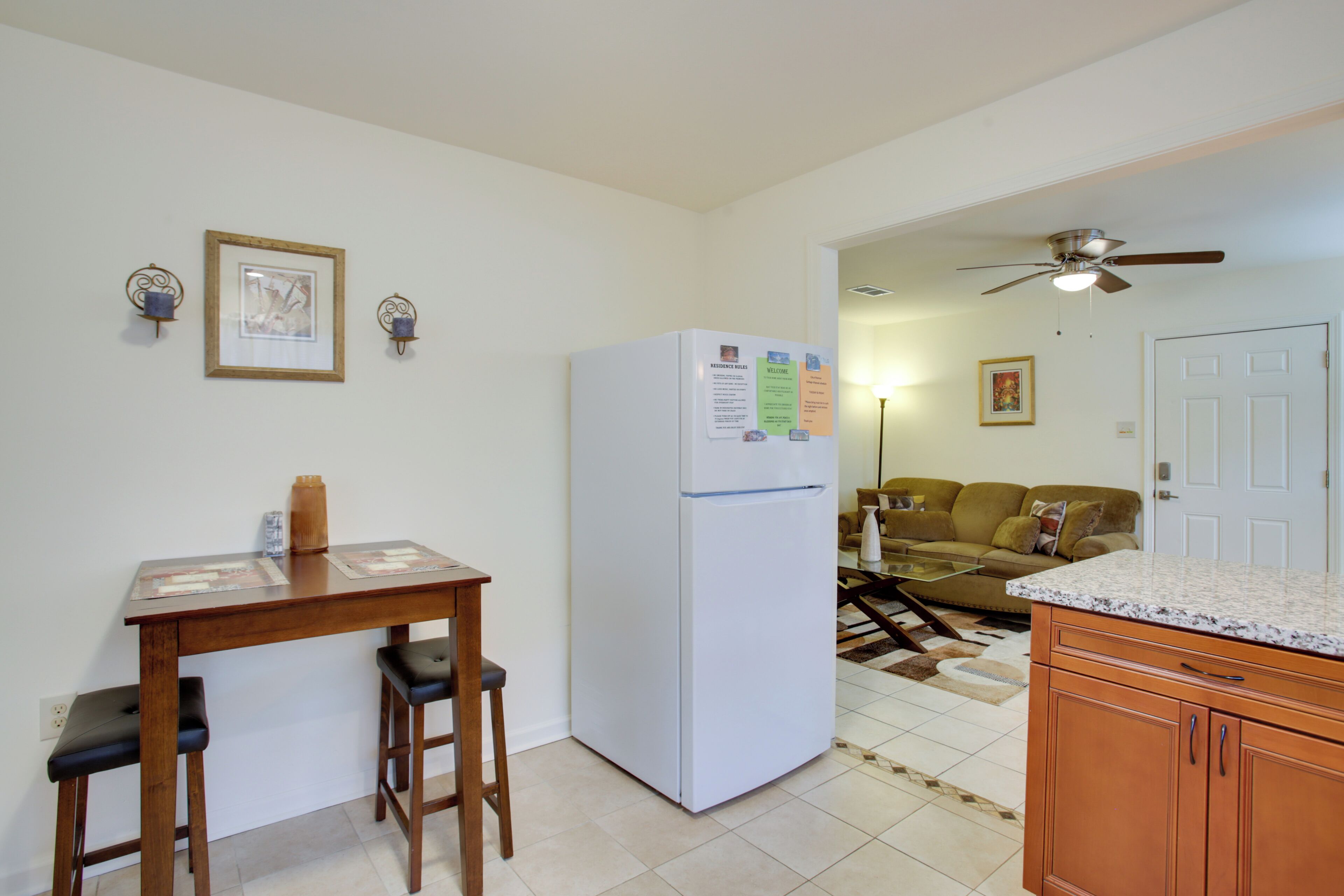 A kitchen with a refrigerator, wooden table, and bar stools. There is a ceiling fan and a door in the background.