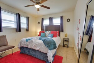 Un dormitorio con cama, una silla, una mesita de noche, ventilador de techo y una ventana con cortinas.