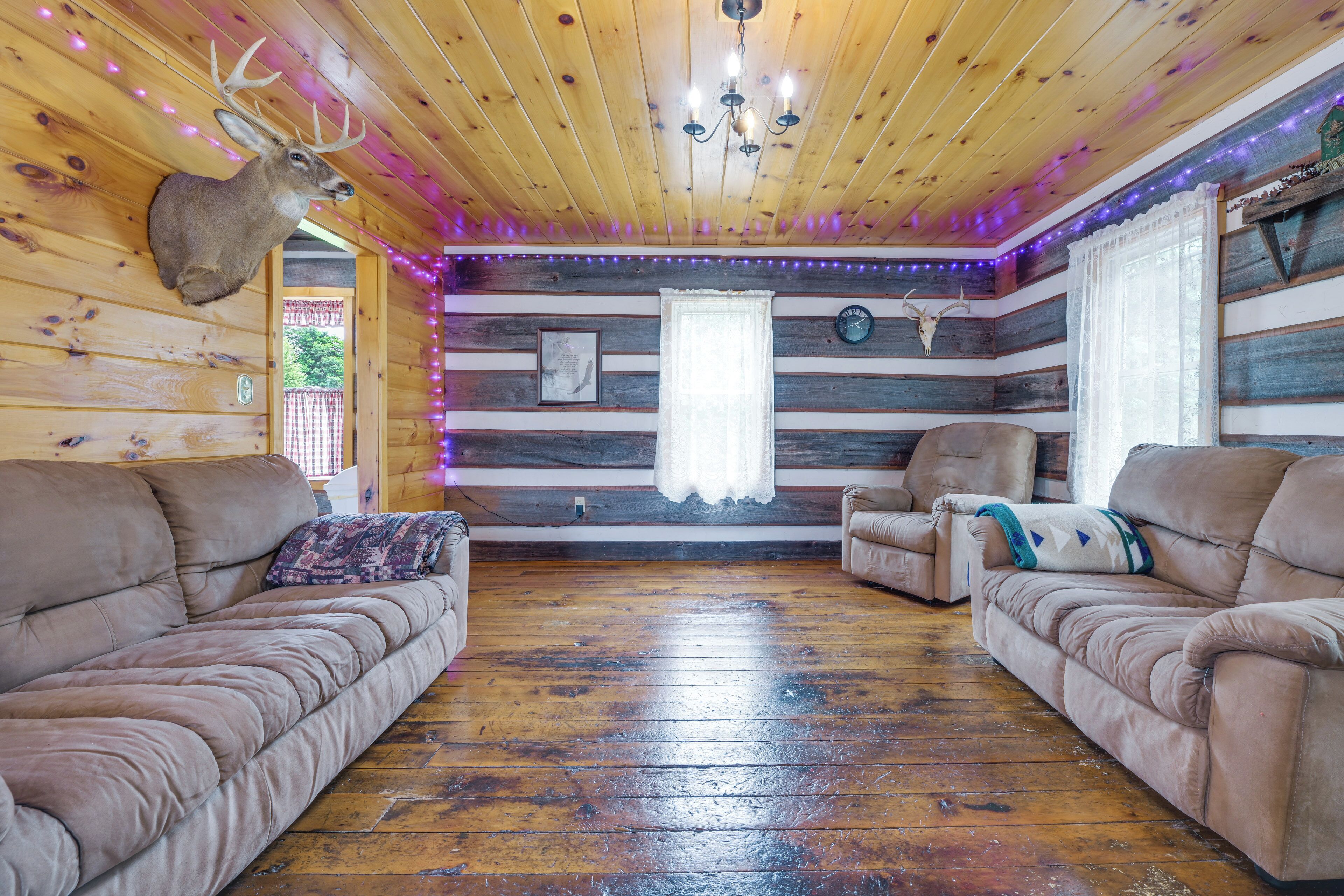 Ein rustikaler Cabin-Innenraum mit Holzwänden, einem aufgehängten Hirschgeweih, zwei Ledersofas, einer Kristallleuchtre und violetten LED-Leuchten.