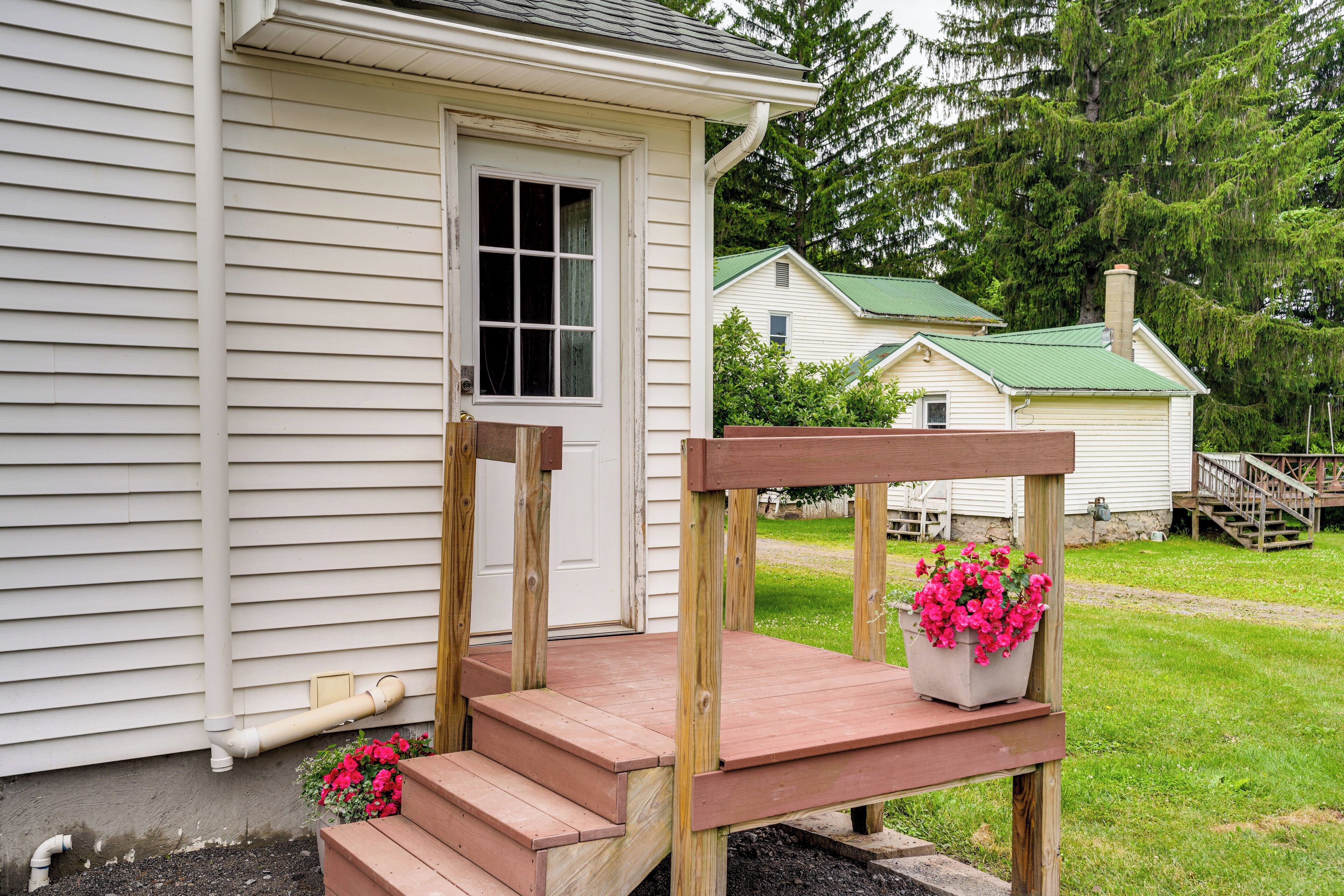 A wooden porch with steps leading to a door, a potted plant with flowers, and a green roofed house in the background.