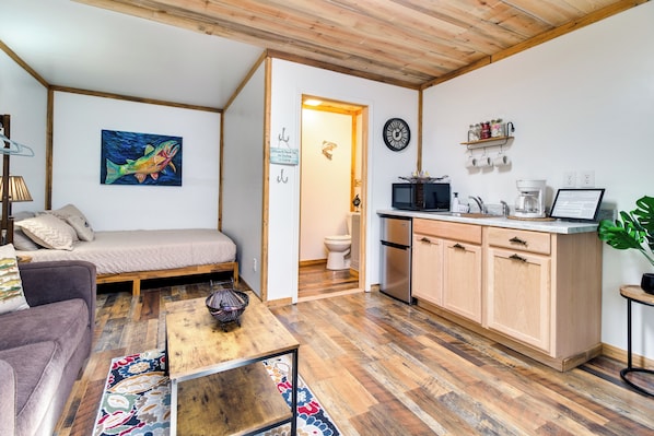A compact living space with a sofa, a coffee table, a painting, a kitchen area with cabinets and appliances, and a wooden ceiling.