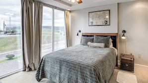 Een slaapkamer met een groot raam, een plafondventilator, een bed met hoofdeinde, een nachtkastje en een wandlamp.