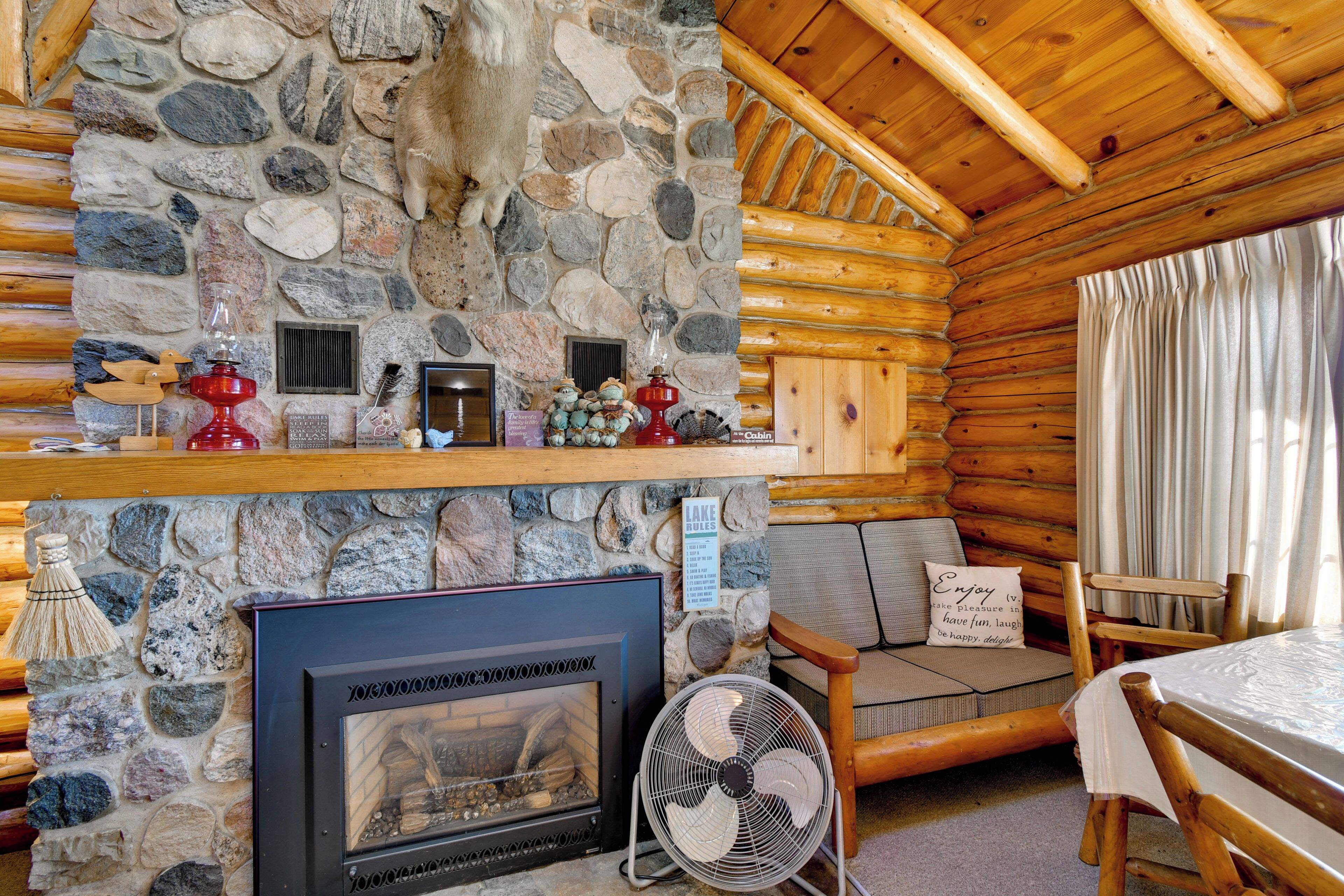 A log cabin interior with a stone fireplace, wooden furniture, and a fan.