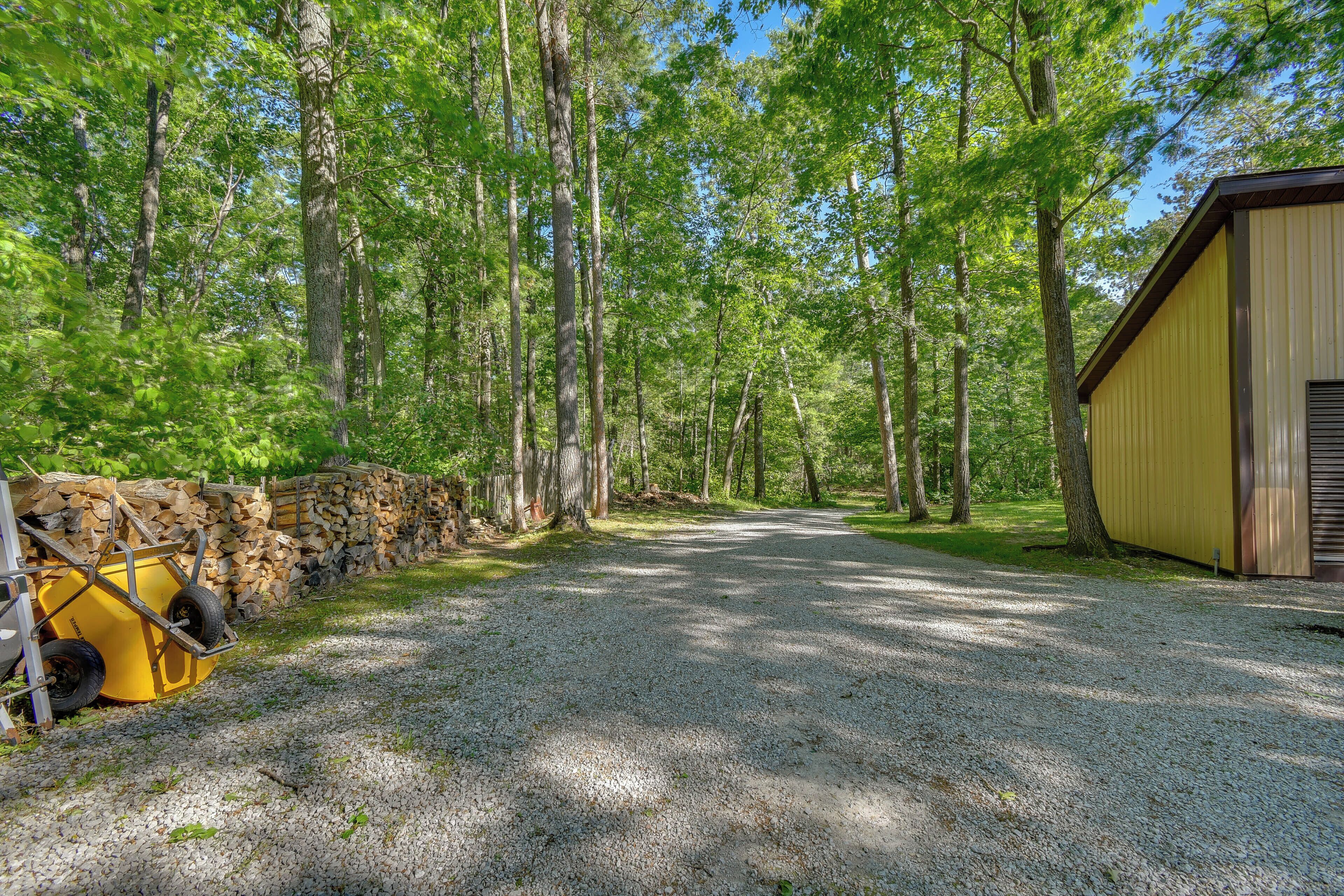 A gravel path leading to a yellow building, surrounded by tall trees and a stack of firewood.