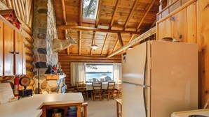 Interior rústico de una cabaña con pared de piedra, techo de madera, comedor y cocina con refrigerador.