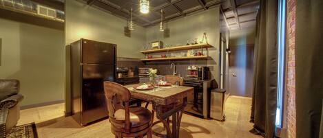 A kitchen with a wooden table, chairs, a refrigerator, and a shelf with kitchenware.