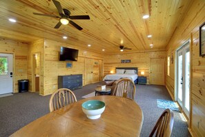 Interior de una cabaña de madera con una mesa de comedor, sillas, un ventilador de techo, un televisor montado en la pared y una cama con cabecera.