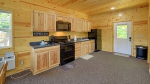 Interior de una cabaña de madera con cocina, que incluye estufa, microondas y refrigerador, un letrero de cafetería y una puerta con ventana.
