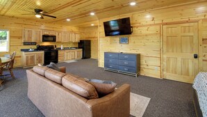 Interior de una cabaña de madera con kitchenette, televisor montado en la pared, sofá y comedor.