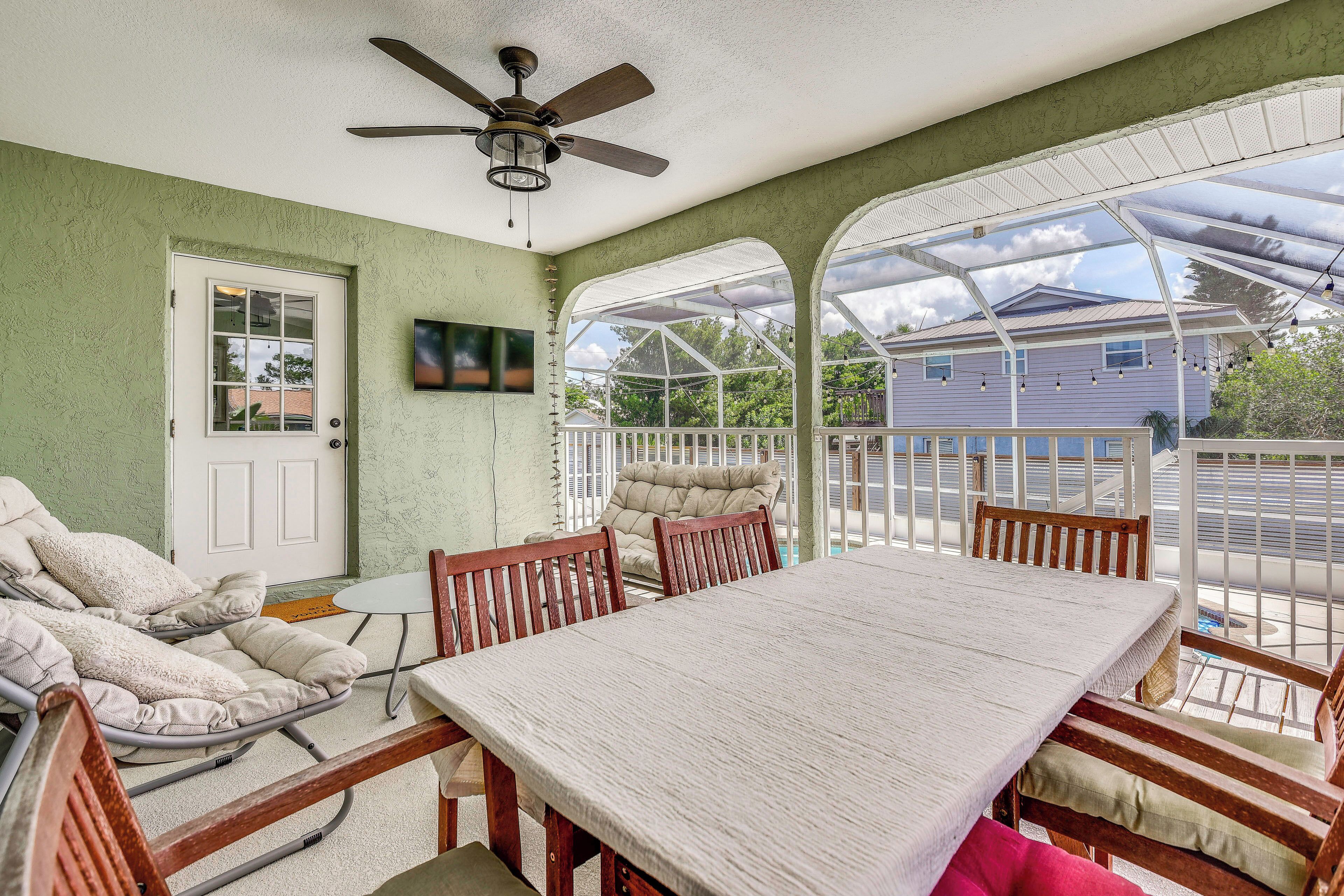 Une terrasse couverte avec une table et des chaises, un ventilateur de plafond et une vue sur une maison à travers la rambarde en verre.
