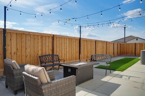 An outdoor patio with wicker furniture, a glass-top table, string lights, and a wooden fence.