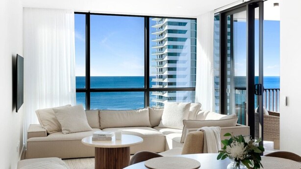 Ein modernes Wohnzimmer mit Sofa, Couchtisch und Essbereich, mit Blick auf ein Hochhaus und das Meer.