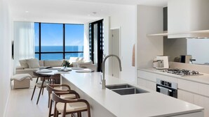 Eine moderne Küche mit einer zentralen Kochinsel, ein Essbereich mit Meerblick und ein Wohnbereich mit Sofa und Sesseln.