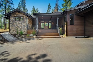 A two-story house with a stone facade, a wooden deck, and a driveway leading to the entrance. - NC407 | 56304 Elk Run Drive (Sunriver)