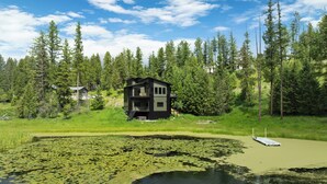 Une cabane moderne à deux étages, entourée d’un étang parsemé de nénuphars, d’une forêt dense et d’un ciel dégagé.